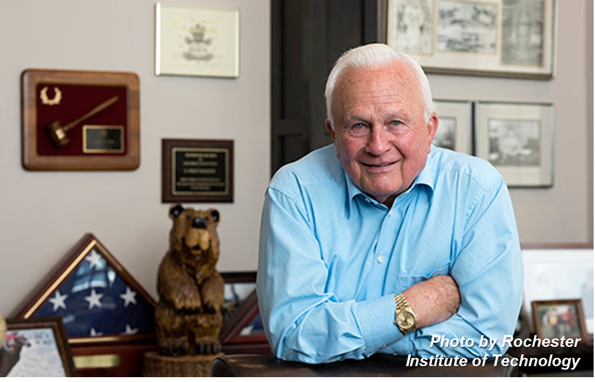 E. Philip Saunders photographed leaning on a wooden desk, with numerous plaques and certificates on the wall behind him.